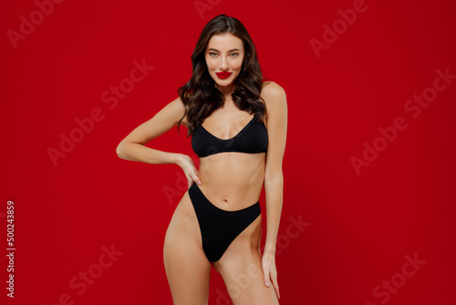 Young stunning sexy brunette woman 20s with perfect fit body wear black underwear stand akimbo arm on waist lok camera isolated on plain red background studio portrait. People female beauty concept