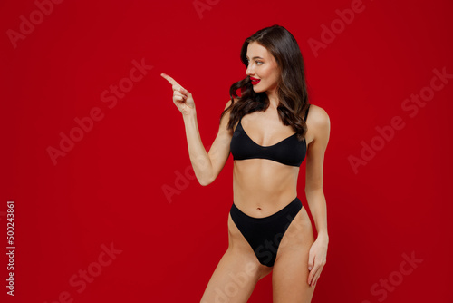 Young sexy hot brunette woman 20s with perfect fit body wear black underwear point index finger aside on workspace area mock up isolated on plain red background studio. People female beauty concept