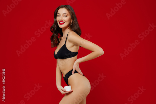 Side view young sexy woman 20s with perfect fit body wear black underwear applying moisturizer, body anti-cellulite cream from container isolated on plain red background. People female beauty concept