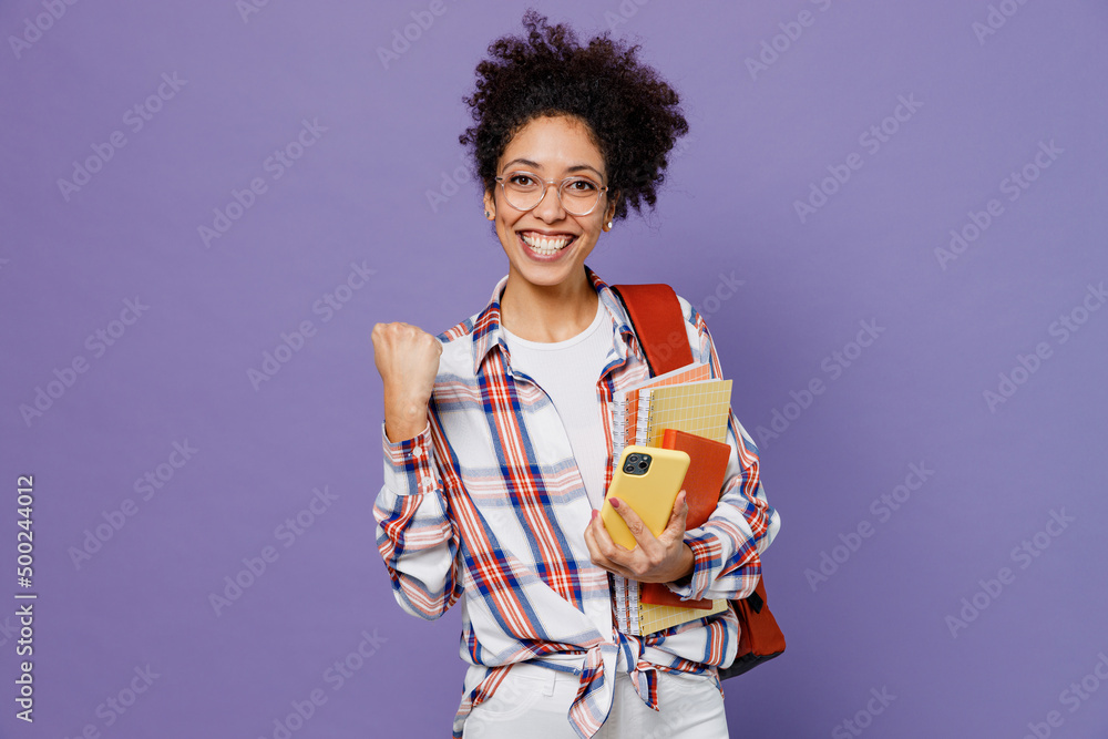 Young fun girl woman of African American ethnicity teen student in shirt backpack use mobile cell phone do winner gesture isolated on plain purple background. Education in high school college concept. Young fun girl woman of African American ethnicity teen student in shirt backpack use mobile cell phone do winner gesture isolated on plain purple background. Education in high school college concept.