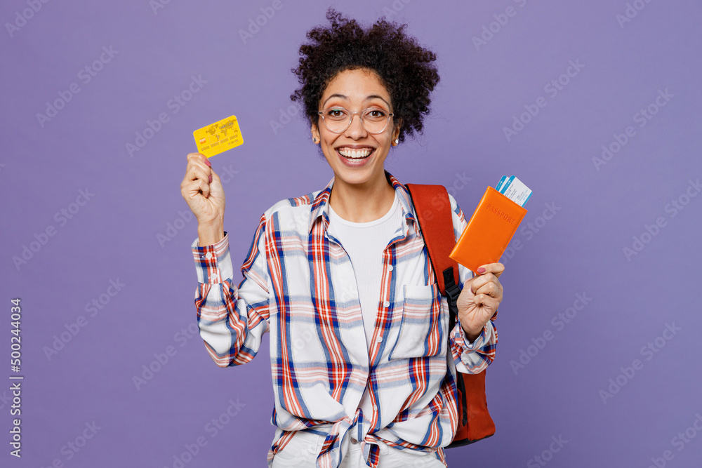 Young happy tourist girl woman of African American ethnicity student in shirt backpack travel abroad hold passport ticket credit card isolated on plain purple background. Air flight journey concept. Young happy tourist girl woman of African American ethnicity student in shirt backpack travel abroad hold passport ticket credit card isolated on plain purple background. Air flight journey concept.