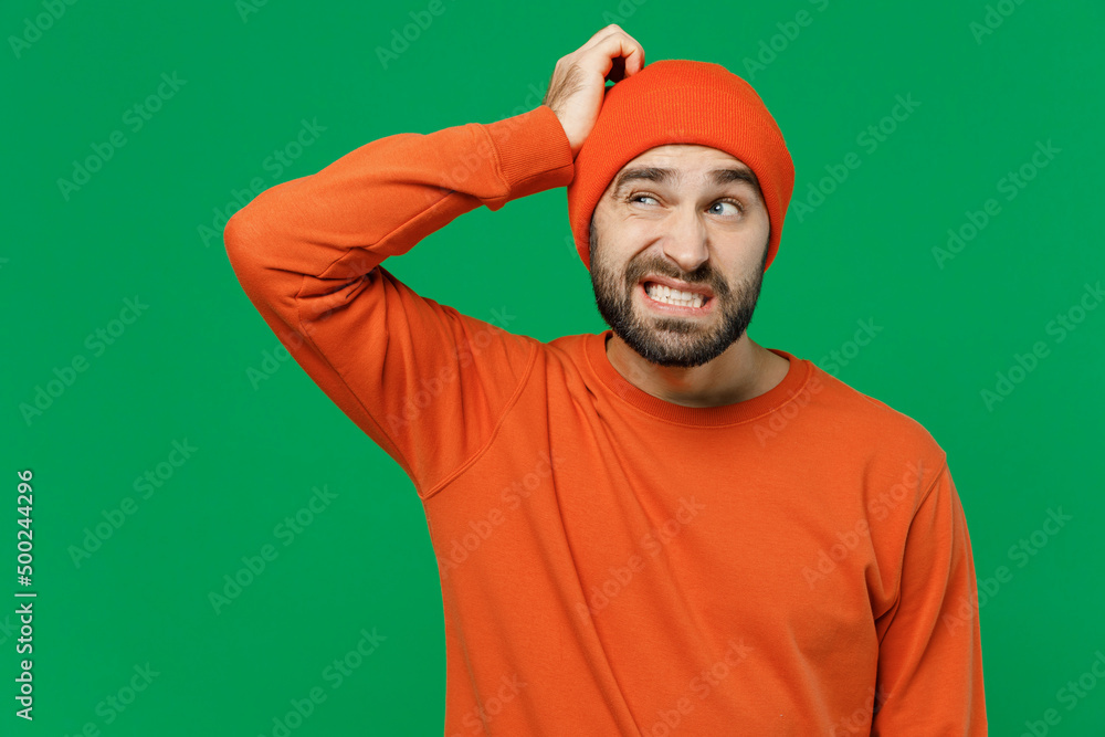 Fototapeta premium Young sad puzzled mistaken confused caucasian man 20s wearing orange sweatshirt hat look aside hold scratch head say oops isolated on plain green background studio portrait. People lifestyle concept.