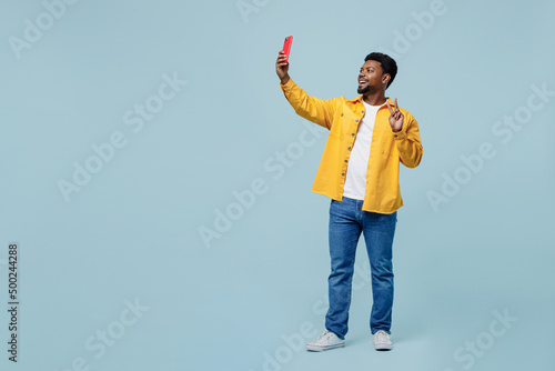 Fotografija Full body fun young man of African American ethnicity wear yellow shirt doing selfie shot on mobile cell phone post photo on social network show v-sign isolated on plain pastel light blue background