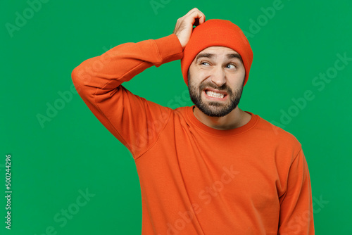 Young sad puzzled mistaken confused caucasian man 20s wearing orange sweatshirt hat look aside hold scratch head say oops isolated on plain green background studio portrait. People lifestyle concept. photo