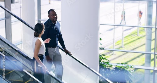Catching up as they head out. Two young businesspeople coming down the escalator at work. Two businesspeople smiling and talking on the escalator at work