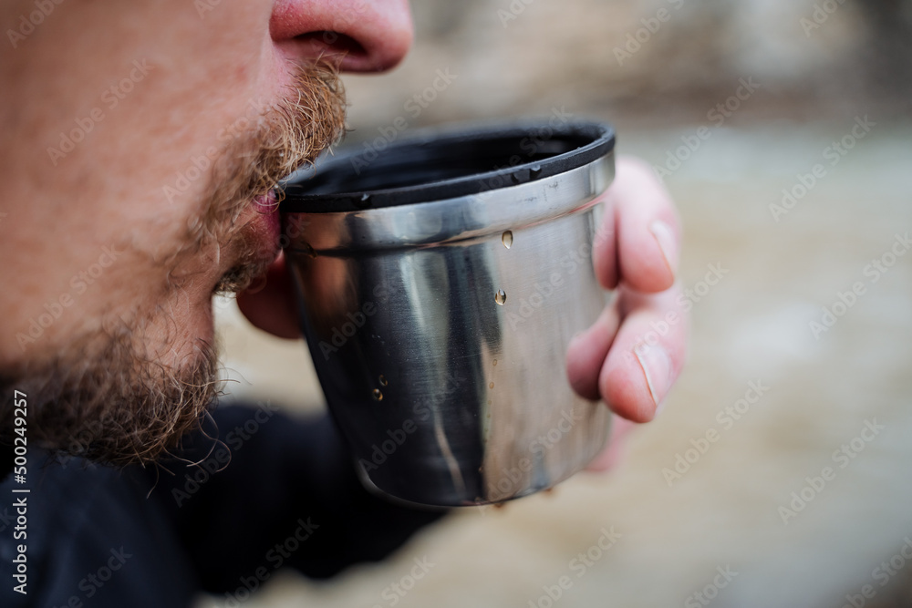 A bearded tourist man drinks hot tea from a mug touching his lips to a glass, bring a cup of coffee to his mouth, drink a drink, breakfast in nature, a tourist lunch. A bearded tourist man drinks hot tea from a mug touching his lips to a glass, bring a cup of coffee to his mouth, drink a drink, breakfast in nature, a tourist lunch.