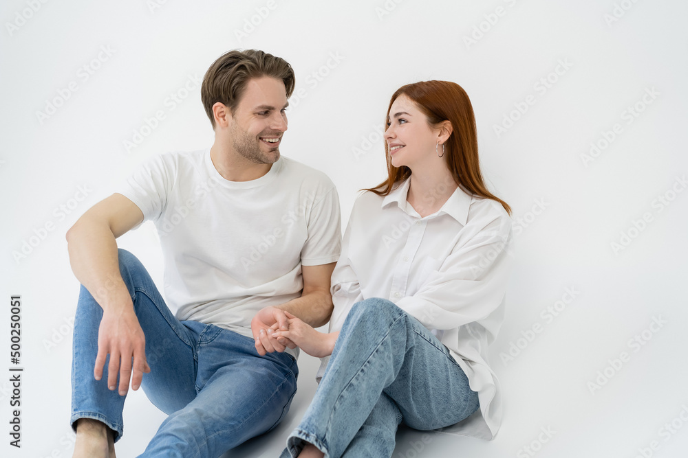 Positive couple in jeans holding hands while sitting on white background. Positive couple in jeans holding hands while sitting on white background.