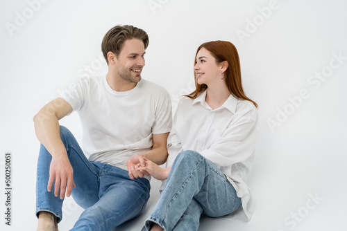 Positive couple in jeans holding hands while sitting on white background.