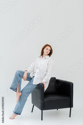 Barefoot woman in jeans sitting on armchair on white background.