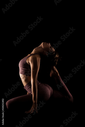 Fit woman practicing yoga poses. Side lit half silhouette girl doing exercise in studio against black background..