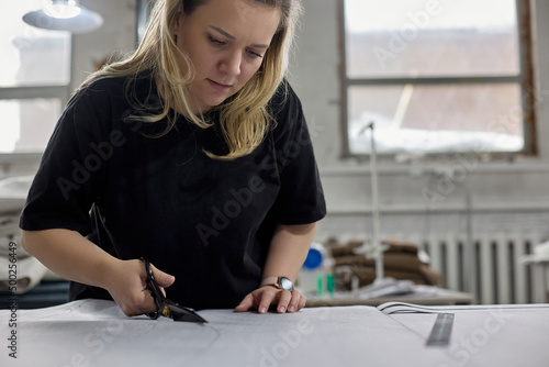 Pretty blonde dressmaker cuts a piece of fabric with scissors in a sewing workshop. Process of making a piece of clothing or upholstery for upholstered furniture from fabric in a studio workshop