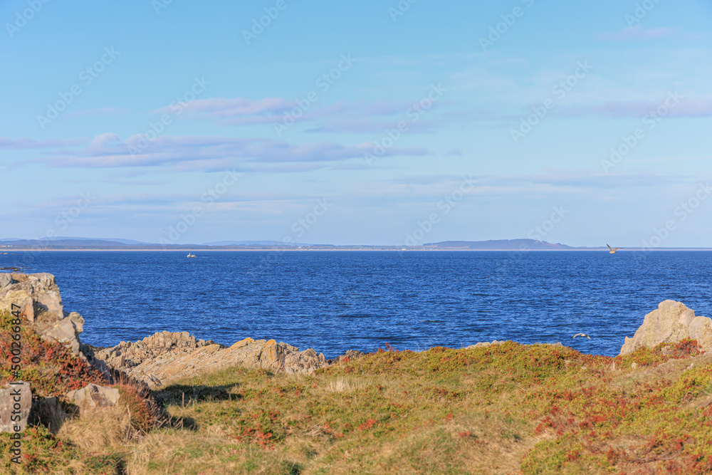 A scenic beautiful view of a Scotish wild seaside with majestic choppy blue sea with mountains summit in the background and grassy beach on the foreground under a beautiful blue sky and white clouds A scenic beautiful view of a Scotish wild seaside with majestic choppy blue sea with mountains summit in the background and grassy beach on the foreground under a beautiful blue sky and white clouds