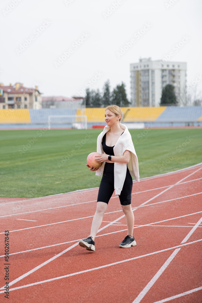 full length view of smiling sportswoman walking on stadium with ball.