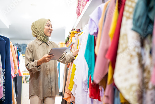 business owner wearing hiijab using a tablet while holding clothes on a hanger at a Muslim clothing store photo