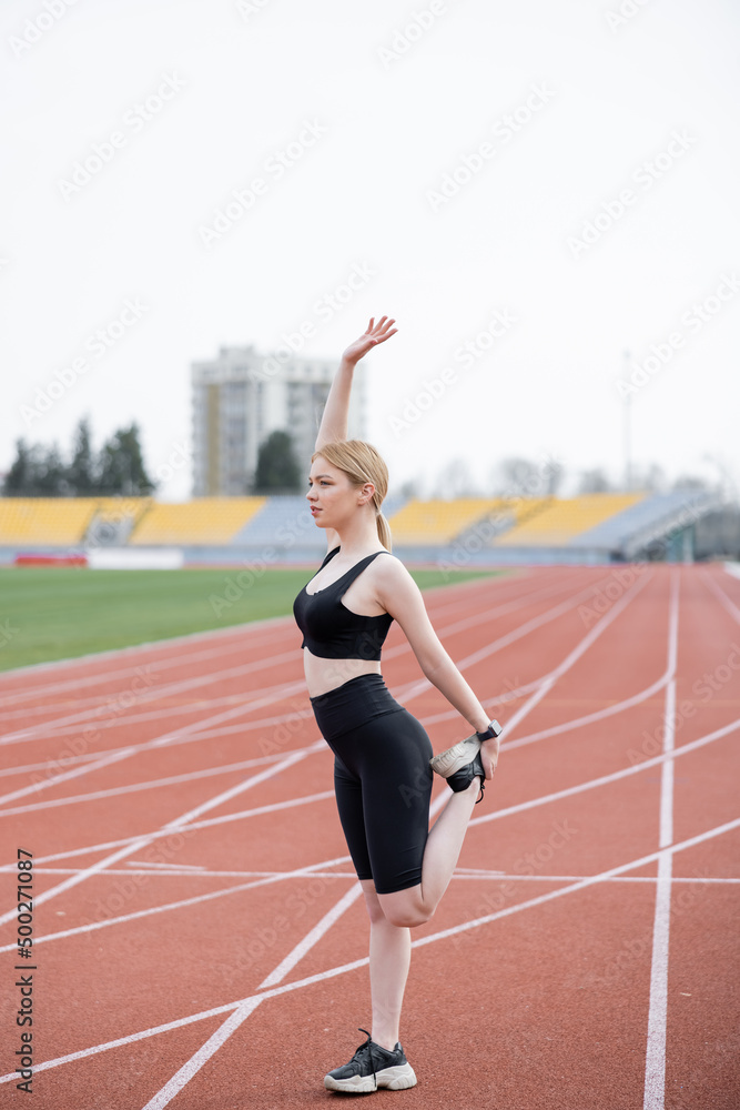 full length view of sportswoman standing with raised hand while stretching on athletic field.