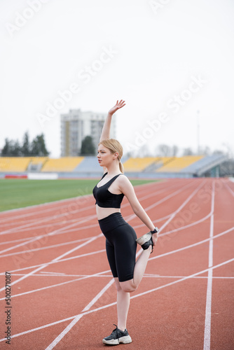 full length view of sportswoman standing with raised hand while stretching on athletic field.