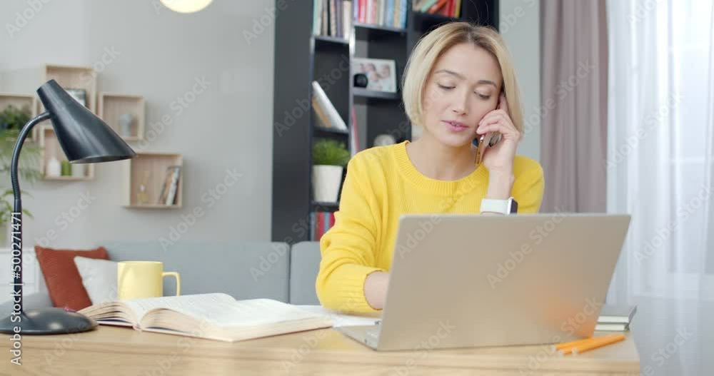 Busy young woman in casual attire typing on laptop and talking on mobile phone while sitting at desk. Working process of business lady at home. Busy young woman in casual attire typing on laptop and talking on mobile phone while sitting at desk. Working process of business lady at home.