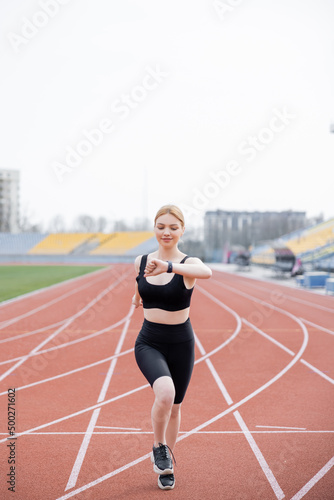 full length view of happy sportswoman running on stadium and looking on fitness tracker.