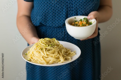 A woman in a blue dress holds a plate of pasta in one hand and a bowl of pasta in the other hand