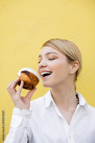 excited woman with closed eyes eating cupcake isolated on yellow.