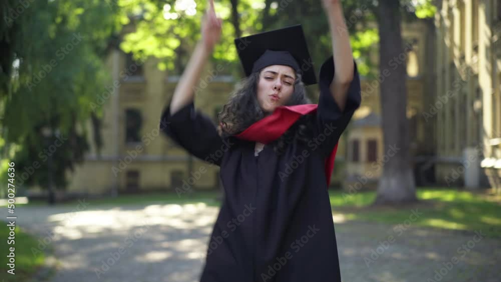 Cheerful young beautiful woman in graduation toga dancing at university campus in the morning looking at camera. Portrait of excited intelligent Caucasian graduate student posing having fun outdoors Cheerful young beautiful woman in graduation toga dancing at university campus in the morning looking at camera. Portrait of excited intelligent Caucasian graduate student posing having fun outdoors