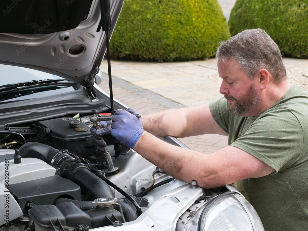 A mechanic wearing blue gloves works in the engine bay of a sports car with a socket set spanner. A mechanic wearing blue gloves works in the engine bay of a sports car with a socket set spanner.