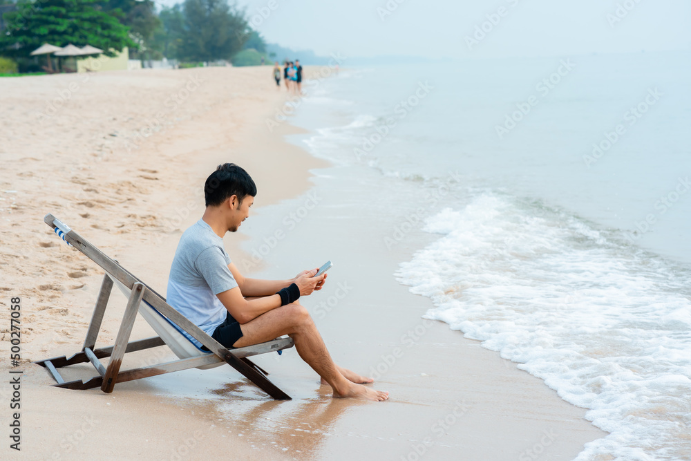 A handsome Asian man sits in a chair on his laptop computer by the sea as the waves crash against the shore, comfortably touching his feet,work from home.