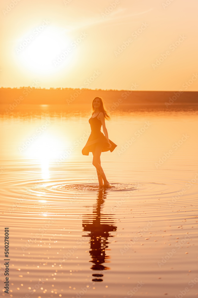 Silhouette of a young woman in an airy black dress in the water of a lake with sky reflection.
