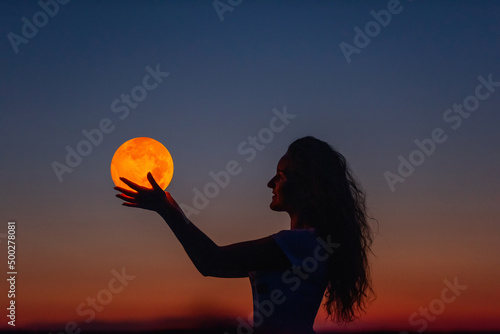 A young woman holds the full moon in her hands against the backdrop of a red sunset. Astrology