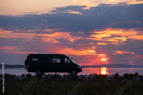 Backlighting silhouette of a camper next to a salt lake at sunset with the sun reflecting in it © JoseMaria