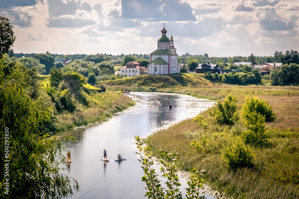 Summer landscape in Suzdal. Church of Elijah the Prophet on the bank of the river Kamenka. Tourists on SUP boards float on the river. Russia, Vladimir region, Golden Ring of Russia
