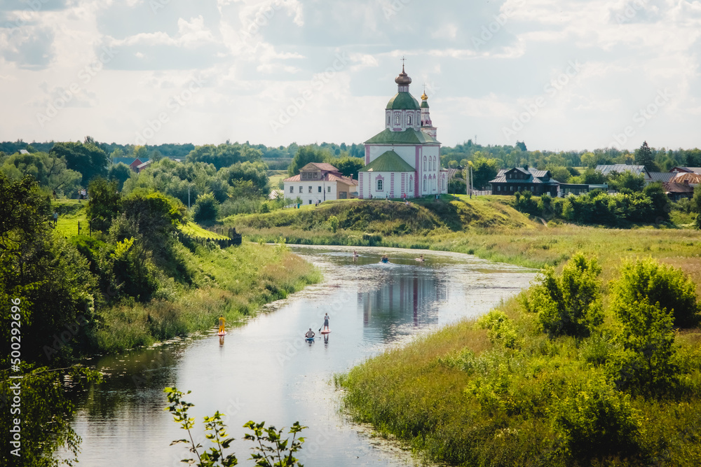 Fototapeta premium Summer landscape in Suzdal. Church of Elijah the Prophet on the bank of the river Kamenka. Tourists on SUP boards float on the river. Russia, Vladimir region, Golden Ring of Russia