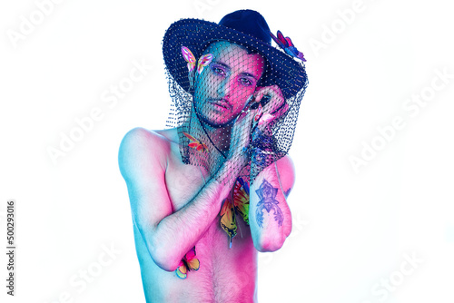 Young model with tattoos, wearing a hat with a mesh, hanging butterflies as decorative accessories and makeup. Posing with white background, magenta and blue light. Showing her feelings.