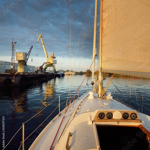 White yacht arriving in port at sunset. Close-up view from the deck to the bow and sails. Dramatic sky after the storm. Baltic sea. Nautical vessel, transportation, service, sport, recreation concepts