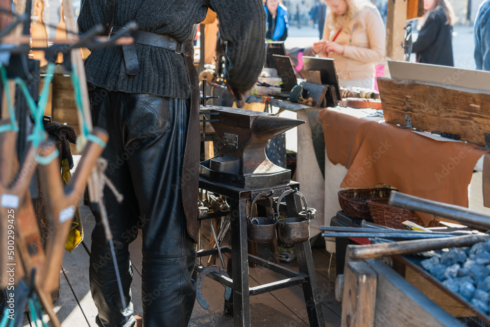 A blacksmith in black leather pants, a black leather apron, a knitted sweater works behind an anvil at a fair, a buyer is standing in front of the counter. Ancient professions, handmade