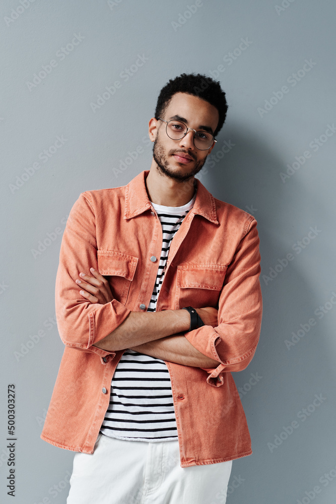 Content successful young Black man in coral shirt standing with crossed arms against gray wall Content successful young Black man in coral shirt standing with crossed arms against gray wall