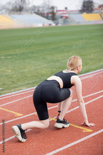 young sportswoman in black bike shorts standing in low start position on athletic field.