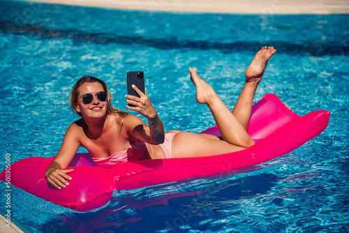 Sexy woman with a phone in a swimsuit lies on a pink inflatable mattress in the pool. Relax by the pool on a hot summer sunny day. Vacation concept. Selective focus
