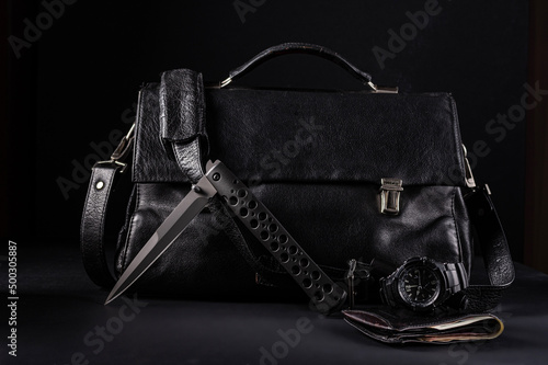 A large black folding knife on the background of a black leather bag. Gentleman's set of knife, watch and wallet. Knife in half-folded position.