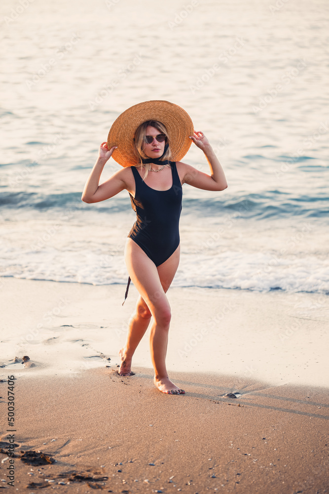 Beautiful girl in a black swimsuit and hat on a sandy beach at the sea in the sunset sunlight