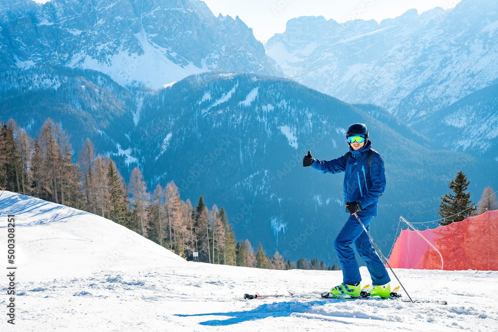 Skier gesturing thumbs up while skiing on snow covered landscape. Scenic view of majestic mountain range against sky. Male tourist enjoying winter sport in alps.
