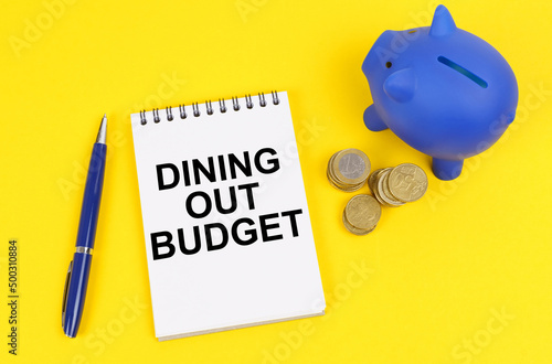 On a yellow surface, a piggy bank, coins and a notepad with the inscription - Dining out budget