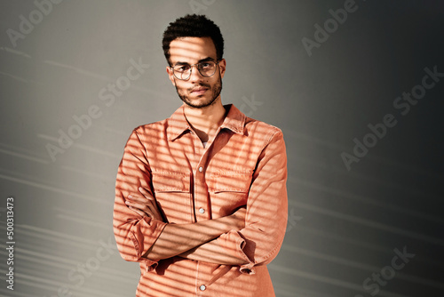 Portrait of serious young black man in coral denim shirt and eyeglasses standing with crossed arms, light through blinds effect