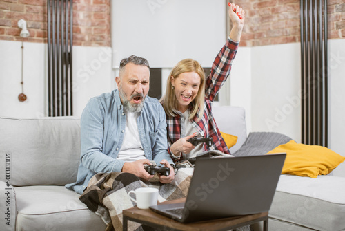 Middle aged caucasian family gesturing emotionally while winning in video games at home. Happy married couple using wireless joysticks and laptop for having fun during free time.