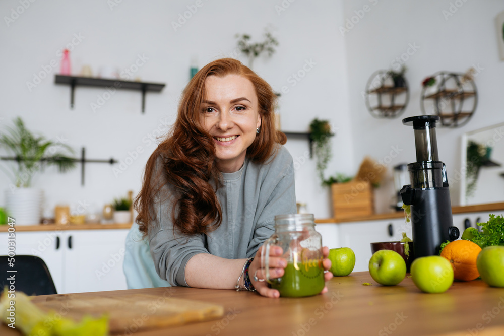 portrait A red-haired woman with long hair made a green smoothie from fresh vegetables and fruits in her bright kitchen