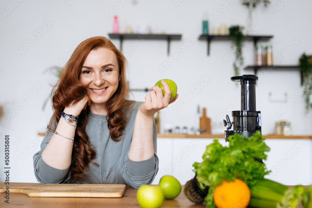 A woman with red hair is sitting at the table in her kitchen smiling, looking at the camera and holding a green apple in her hands A woman with red hair is sitting at the table in her kitchen smiling, looking at the camera and holding a green apple in her hands