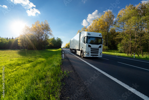 White truck driving on the asphalt road between the forest and the meadow in springtime landscape at sunset