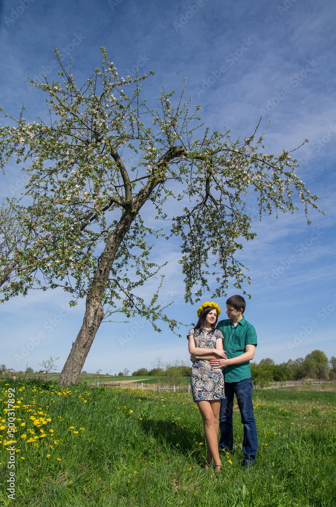Loving couple near a blossoming tree on a background of spring blue sky. Girl with a wreath of dandelions on her head.