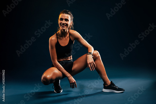 Woman in sport clothes sitting on black background after training. Smiling runner relaxing after intense workout.