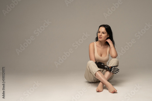 Fotografie Caucasian disabled Woman with amputated hand, artificial arm sitting looking to left isolated on beige background